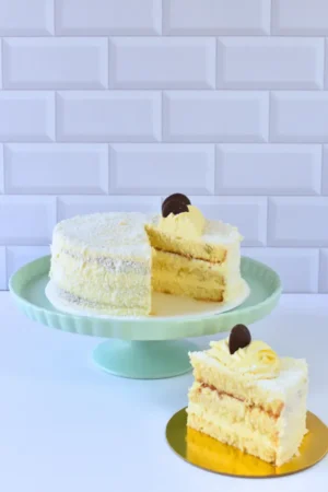 Torta Noche Buena de Pepos Cake Medellín, cubierta en coco rallado y rellena con crema suave, decorada con rosetón amarillo y chocolate, presentada en un pedestal con una porción servida al lado.