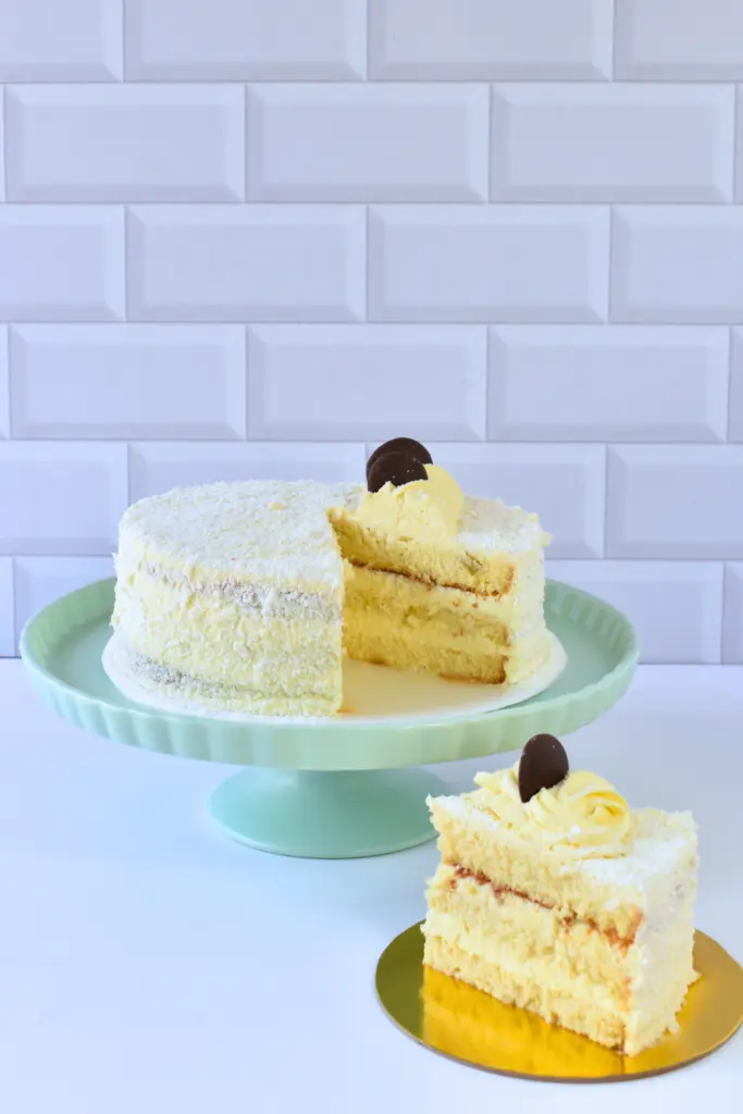 Torta Noche Buena de Pepos Cake Medellín, cubierta en coco rallado y rellena con crema suave, decorada con rosetón amarillo y chocolate, presentada en un pedestal con una porción servida al lado.