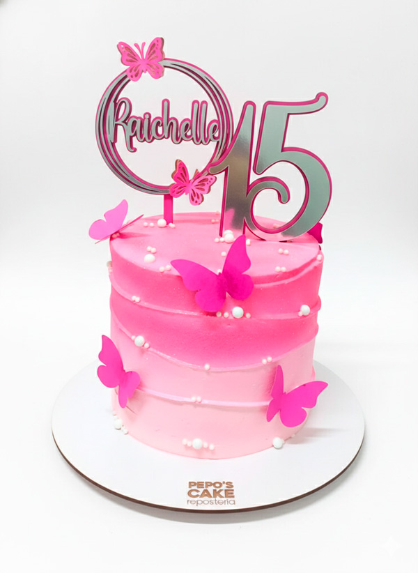 Torta de cumpleaños rosa decorada con mariposas sobre un fondo blanco limpio. El pastel presenta un degradado de color rosa suave a fucsia, adornado con pequeñas perlas blancas comestibles. En la parte superior tiene un topper plateado personalizado con el nombre "Raichelle" y el número "15", decorado con mariposas a juego. La torta reposa sobre una base blanca que incluye el logotipo de "Pepo's Cake Repostería"